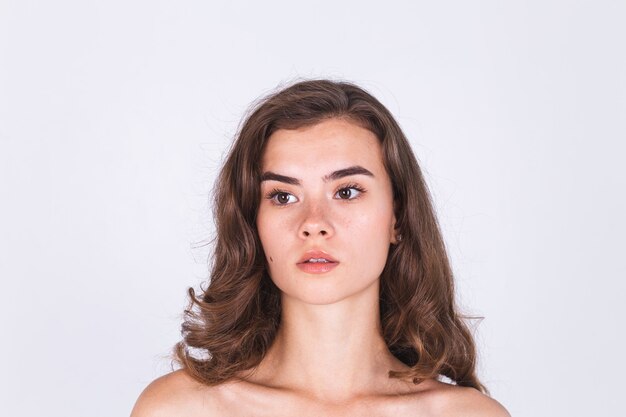 Jeune belle femme naturelle à la peau douce et propre avec un maquillage léger de taches de rousseur sur un mur blanc avec les épaules nues