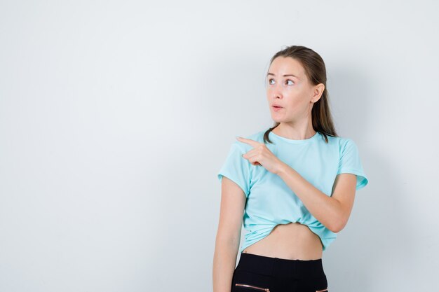 Jeune belle femme pointant vers le coin supérieur gauche, regardant loin en t-shirt et pensif. vue de face.