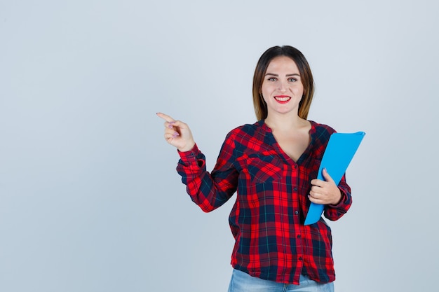 Jeune belle femme pointant vers le coin supérieur gauche, tenant un dossier dans une chemise décontractée, un jean et l'air joyeux, vue de face.