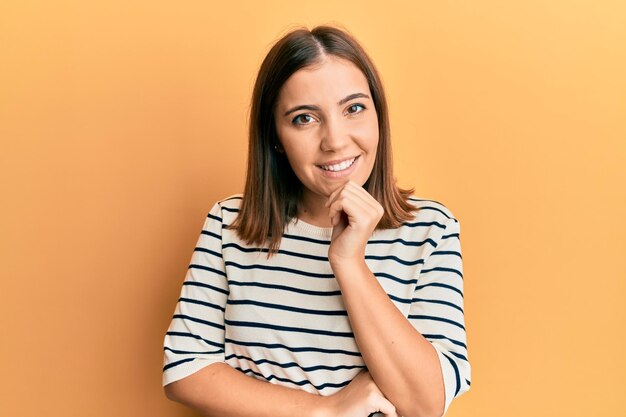 Jeune belle femme portant un t-shirt rayé décontracté souriant regardant confiant à la caméra avec les bras croisés et la main sur le menton pensant positif