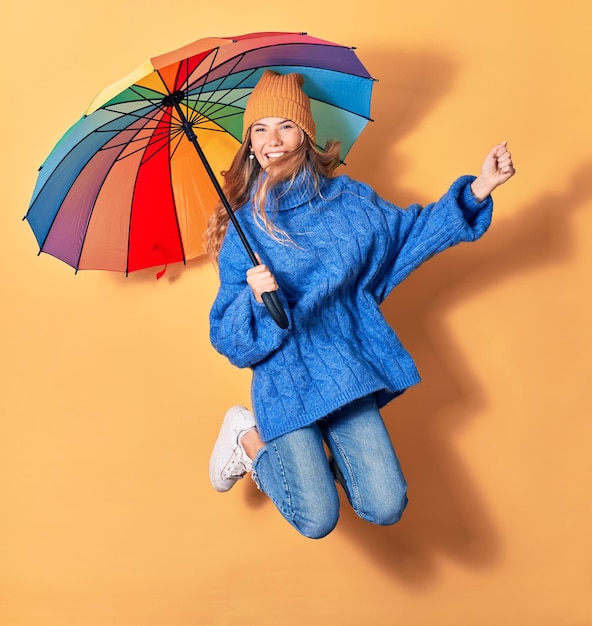 Photo gratuite jeune belle femme portant des vêtements d'hiver souriant heureux sautant avec le sourire sur le visage tenant un parapluie coloré sur fond jaune isolé