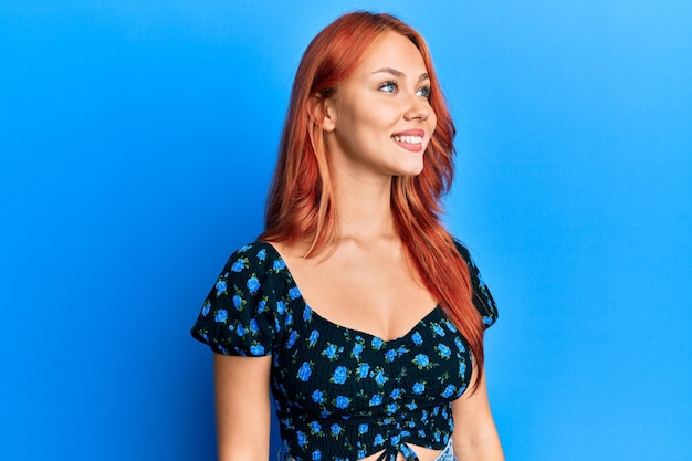 Jeune belle femme rousse portant des vêtements décontractés sur fond bleu regardant de côté avec le sourire sur le visage expression naturelle rire confiant