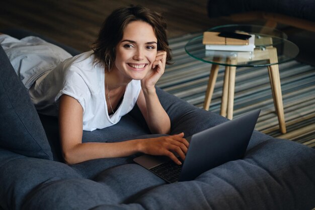 Jeune belle femme souriante allongée sur un canapé avec un ordinateur portable regardant joyeusement à huis clos dans un bureau moderne
