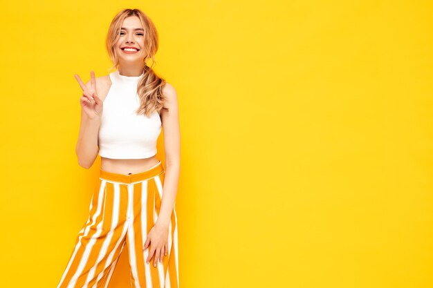 Jeune belle femme souriante dans des vêtements d'été à la mode Sexy femme insouciante posant près de bleu jaune en studio Modèle positif s'amusant Joyeux et heureux Montre le signe de la paix