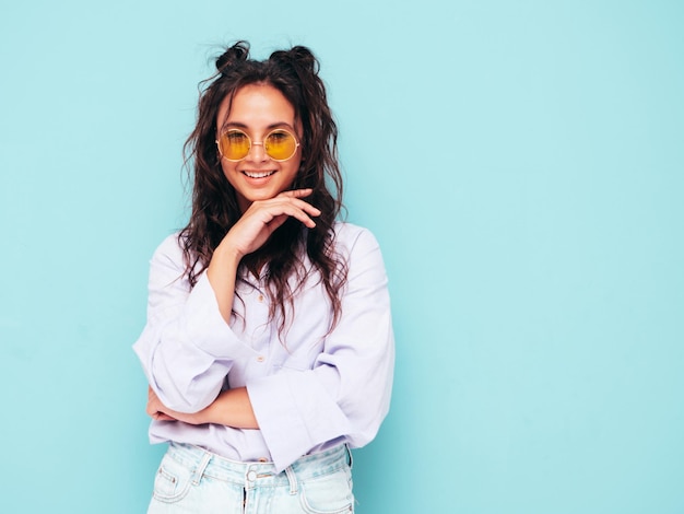 Jeune belle femme souriante dans des vêtements d'été à la mode Sexy femme insouciante posant près du mur bleu en studio Modèle brune positive s'amusant et devenant fou Gai et heureux en lunettes de soleil