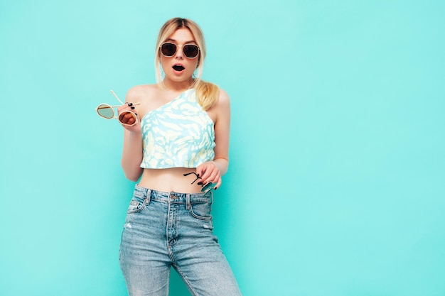 Jeune belle femme souriante dans des vêtements d'été à la mode Sexy femme insouciante posant près du mur bleu en studio Modèle positif tenant de nombreuses lunettes de soleilGai et heureux Montrant et faisant le choix