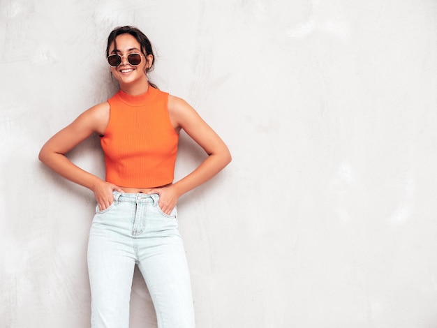 Jeune belle femme souriante dans des vêtements d'été à la mode Sexy femme insouciante posant près d'un mur gris en studio Modèle brune positive s'amuser et devenir fou Gai et heureux en lunettes de soleil