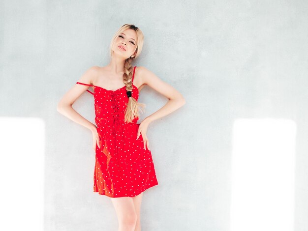 Jeune belle femme souriante en robe rouge d'été à la mode Sexy femme blonde insouciante posant près du mur en studio Modèle positif s'amusant Enthousiaste et heureux à la journée ensoleillée