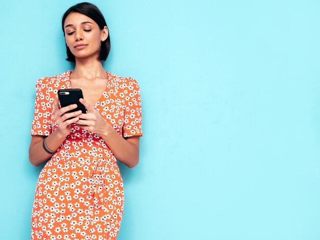 Jeune belle femme souriante en robe rouge d'été à la mode Sexy femme insouciante posant près du mur bleu en studio Modèle regardant l'écran du téléphone portable Tenant le smartphone et utilisant des applications