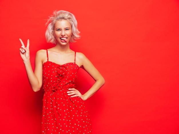 Jeune belle femme souriante en robe rouge d'été à la mode Sexy femme insouciante posant près du mur en studio Modèle blond positif s'amusant et devenant fou Enthousiaste et heureux Montre le signe de la paix