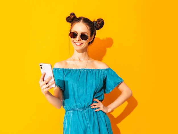 Jeune belle femme souriante en salopette de jeans d'été à la mode Femme insouciante sexy avec une coiffure à deux cornes posant près d'un mur jaune en regardant l'écran du téléphone portable