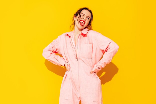 Jeune belle femme souriante en salopette rose d'été à la mode Sexy femme insouciante posant près d'un mur jaune en studio Modèle positif s'amusant et devenant fou Enthousiaste et heureux Montre la langue