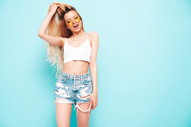 Jeune belle femme souriante en short jeans d'été à la mode Sexy femme blonde insouciante posant près du mur bleu en studio Modèle positif s'amusant Gai et heureux en lunettes de soleil Isolé