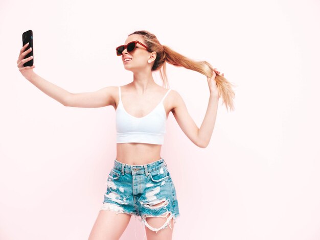 Jeune belle femme souriante en short jeans d'été à la mode Sexy femme blonde insouciante posant près du mur en studio Modèle positif s'amuser Gai et heureux Prendre selfie
