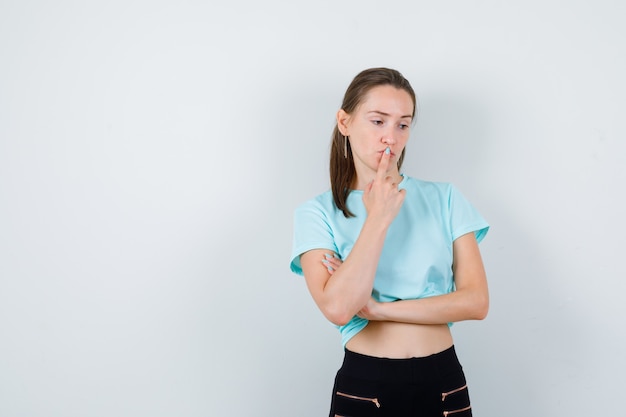 Jeune belle femme en t-shirt, pantalon avec le doigt sur la bouche et l'air pensif, vue de face.