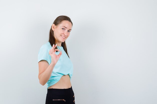 Jeune belle femme en t-shirt, pantalon montrant un geste correct et semblant heureux, vue de face.