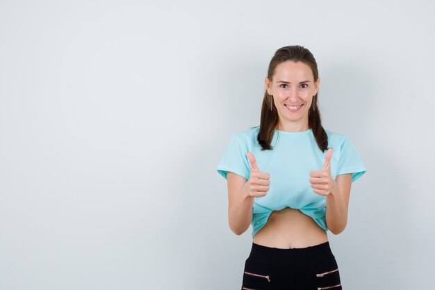 Jeune belle femme en t-shirt, pantalon montrant les pouces vers le haut et l'air joyeux, vue de face.