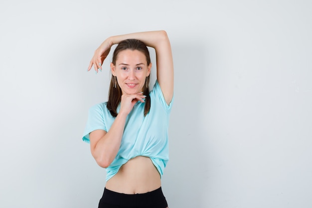 Jeune Belle Femme En T-shirt Posant Tout En Gardant Le Bras Sur La Tête, Le Menton à Portée De Main Et L'air Joyeux, Vue De Face.