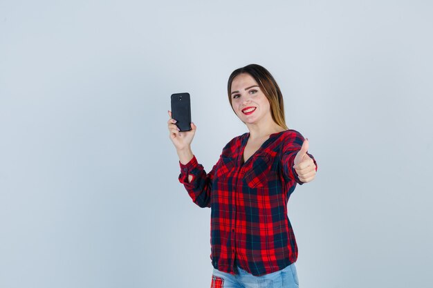 Jeune belle femme tenant un téléphone, montrant le pouce en chemise décontractée et l'air joyeux, vue de face.