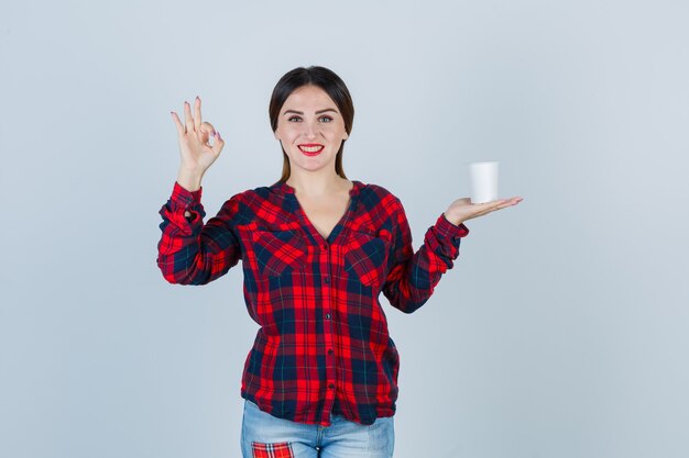Jeune belle femme tenant un verre en plastique, montrant un signe ok en chemise décontractée, un jean et l'air heureux. vue de face.