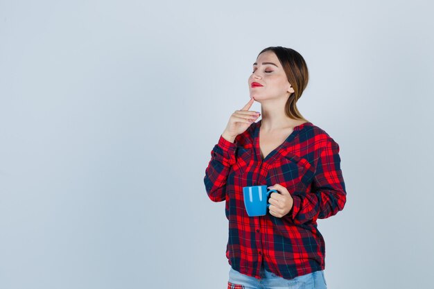 Jeune belle femme touchant le menton avec le doigt tout en tenant une tasse en chemise décontractée, un jean et l'air joyeux. vue de face.