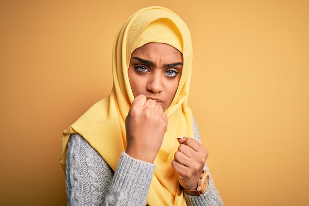 Photo gratuite jeune belle fille afro-américaine portant le hijab musulman sur fond jaune isolé prêt à se battre avec le geste de défense du poing visage en colère et bouleversé peur du problème