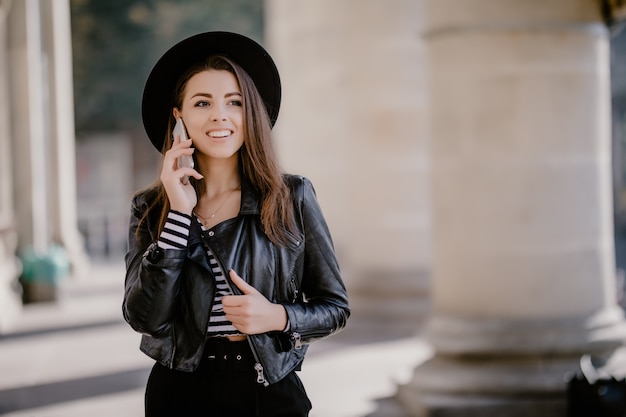 Jeune belle fille aux cheveux bruns dans une veste en cuir, chapeau noir sur la promenade de la ville avoir une conversation téléphonique