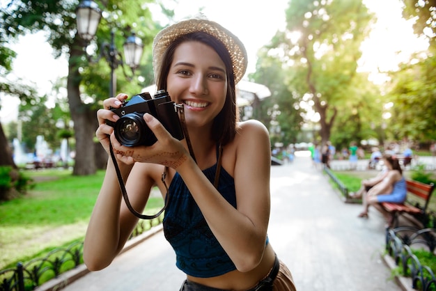 Jeune belle fille brune au chapeau souriant, prenant des photos dans le parc.
