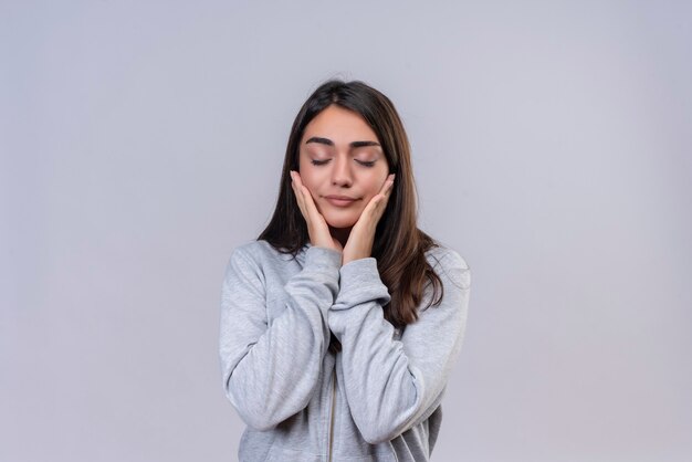 Jeune belle fille à capuche gris à la confiance avec les yeux fermés touchant les joues debout sur fond blanc