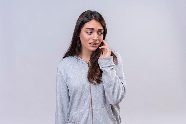 Jeune belle fille à capuche gris avec la main sur le menton en détournant les yeux avec une émotion inquiète debout sur fond blanc