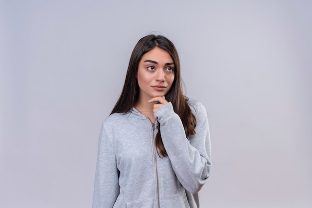 Jeune belle fille à capuche gris à la recherche de suite avec la main sur le menton pensant debout sur fond blanc