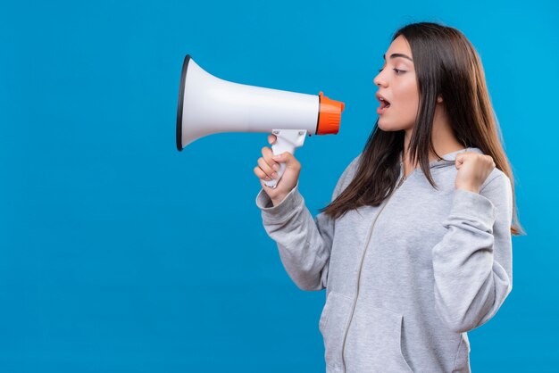 Jeune belle fille à capuche gris à la recherche de suite et parlant au haut-parleur debout sur fond bleu