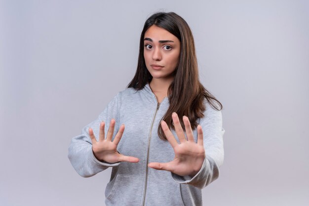 Jeune belle fille à capuche gris regardant la caméra expression agitée faisant le geste d'arrêt debout sur fond blanc