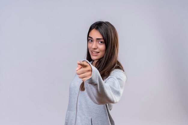 Jeune belle fille à capuche gris regardant la caméra avec une expression insatisfaite et pointant vers la caméra debout sur fond blanc