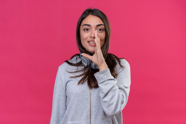 Jeune belle fille à capuche gris regardant la caméra avec le sourire sur le visage main près de la bouche debout sur fond rose