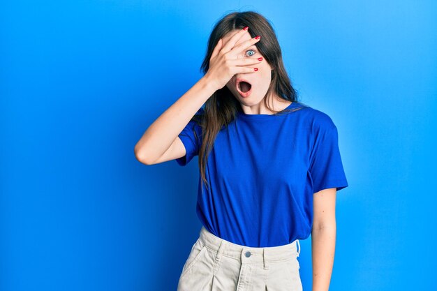 Jeune belle fille caucasienne portant des vêtements décontractés furtivement sous le choc couvrant le visage et les yeux avec la main regardant à travers les doigts effrayés