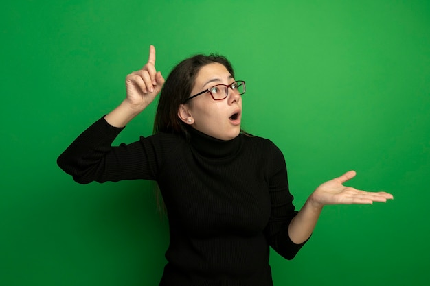 Jeune belle fille dans un col roulé noir et des lunettes à côté surpris et étonné montrant l'index figner présentant quelque chose avec bras