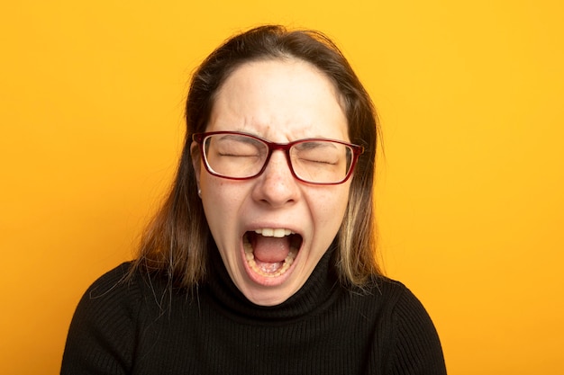 Photo gratuite jeune belle fille dans un col roulé noir et des lunettes criant avec une expression agacée se déchaînant