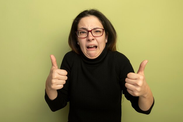 Jeune belle fille dans un col roulé noir et des lunettes étant confus montrant les pouces vers le haut