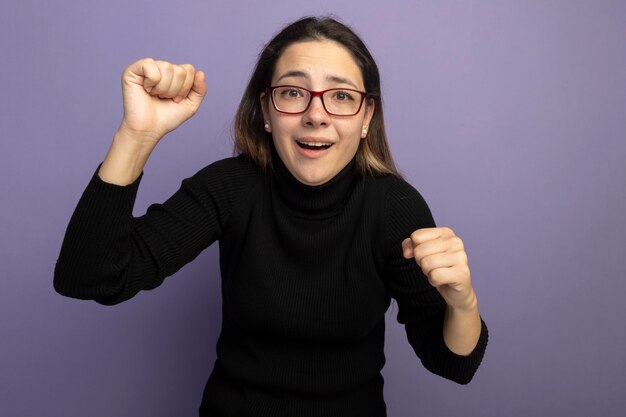 Jeune belle fille dans un col roulé noir et des lunettes serrant les poings souriant étant confus