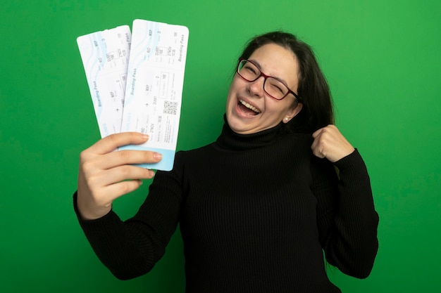 Jeune Belle Fille Dans Un Col Roulé Noir Et Des Lunettes Tenant Des Billets D'avion Heureux Et Excité Poing Serrant