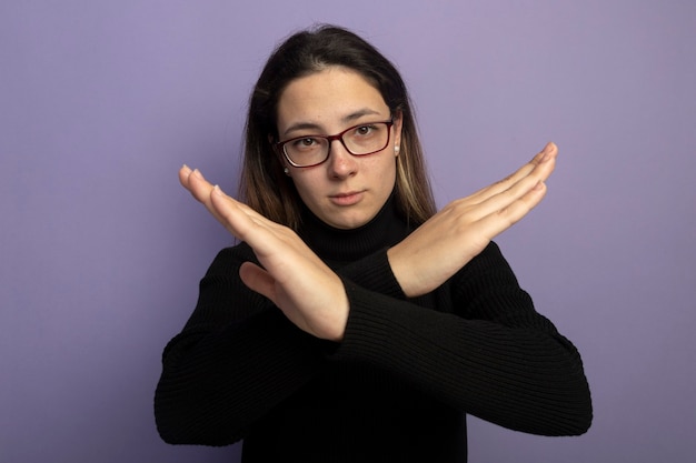 Jeune belle fille dans un col roulé noir et des lunettes avec un visage sérieux faisant le geste stp croisant les mains