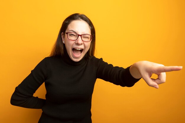 Jeune belle fille dans un pointign à col roulé noir avec l'index à quelque chose en criant excité et heureux