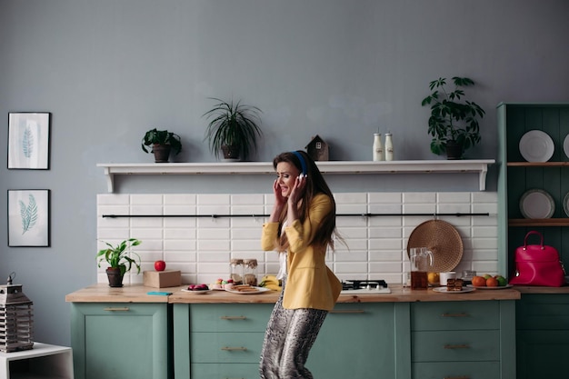 Jeune belle fille dans des vêtements à la mode s'amusant sur la cuisine Heureux modèle brune aux cheveux longs écoutant de la musique et dansant Femme élégante en veste jaune et casque bleu chantant