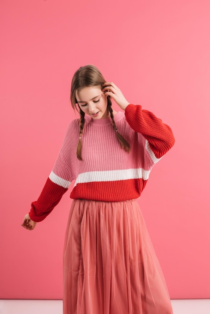 Jeune belle fille avec deux tresses en pull et jupe longue tenant la main près de la tête tout en dansant rêveusement passer du temps sur fond rose