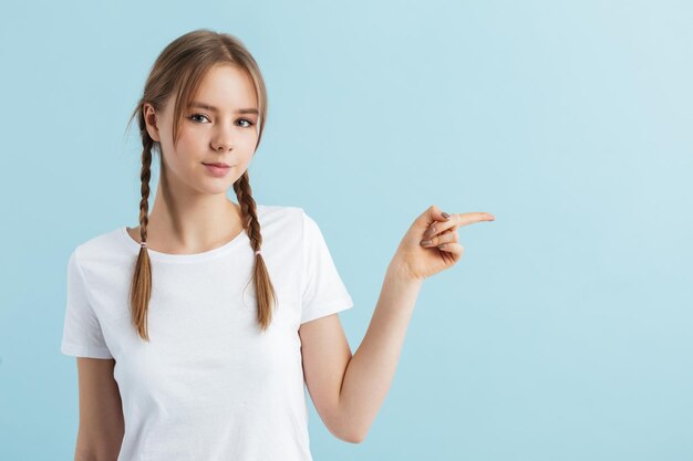 Jeune belle fille avec deux tresses en t-shirt blanc regardant rêveusement à huis clos montrant avec l'index à l'espace vide sur fond bleu isolé