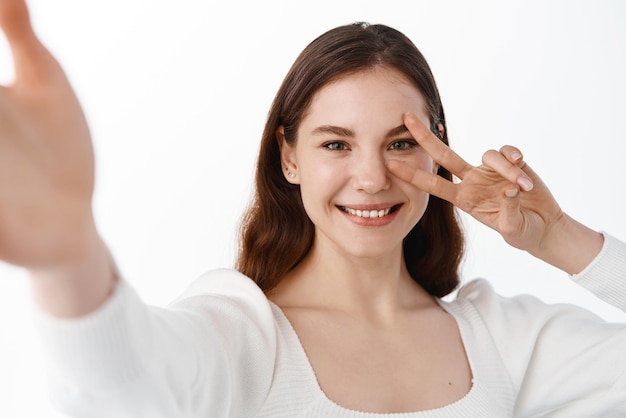 Jeune belle fille étudiante prenant selfie sur appareil photo mobile tenant le téléphone avec la main tendue montrant vsign et souriant debout sur fond blanc