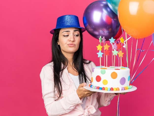 Jeune belle fille mécontente portant un chapeau de fête tenant des ballons avec un gâteau isolé sur un mur rose