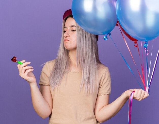 Jeune belle fille mécontente portant un chapeau de fête tenant des ballons et regardant un sifflet de fête dans sa main isolée sur un mur bleu