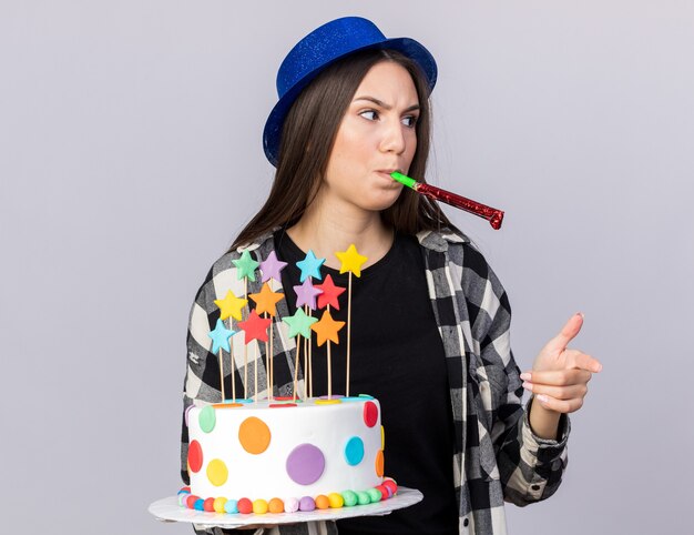 Jeune belle fille mécontente portant un chapeau de fête tenant des points de gâteau soufflant un sifflet de fête isolé sur un mur blanc